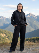 Load image into Gallery viewer, Person wearing a black hoodie and pants standing in front of mountainous landscape